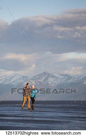 Stock Photograph - A young couple walking on a beach and holding hands with a mountain range in the distance; Anchorage, Alaska, United States of America. Fotosearch