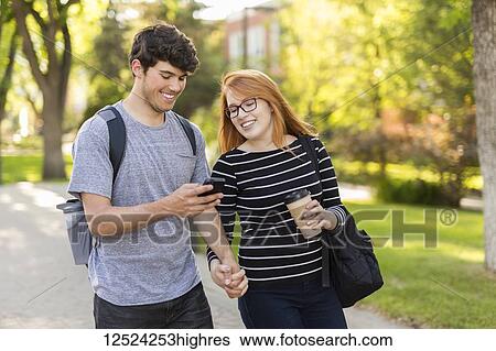Stock Photograph - A young couple walking together and checking social media on a smart phone while on a university campus; Edmonton, Alberta, Canada. Fotosearch