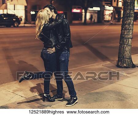 Stock Photograph - A young couple wearing black leather jackets and the woman is lifting one leg while they are standing and kissing on a sidewalk at night; Edmonton, Alberta, Canada. Fotosearch