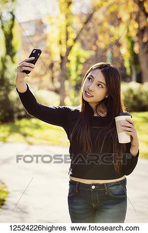 A young female International university student stands holding a coffee cup and poses for a self-portrait with her smart phone; Edmonton, Alberta, Canada View Large Photo Image Stock Photograph - A young female International university student stands holding a coffee cup and poses for a self-portrait with her smart phone; Edmonton, Alberta, Canada. Fotosearch