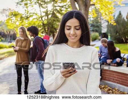 Stock Photograph - A young female university student of Lebanese ethnicity checks her smart phone while her friends are talking together in the background on the university campus; Edmonton, Alberta, Canada. Fotosearch