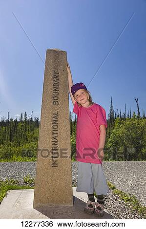 Stock Photograph - A Young Girl poses with the International Boundary marker, United States and Canadian Boarder, Northern Yukon Territory, Canada, Summer. Fotosearch