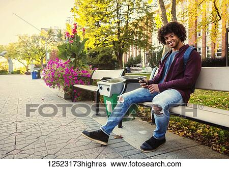 A young male African American university student sits on a bench with his smart phone on a university campus; Edmonton, Alberta, Canada View Large Photo Image Stock Photograph - A young male African American university student sits on a bench with his smart phone on a university campus; Edmonton, Alberta, Canada. Fotosearch