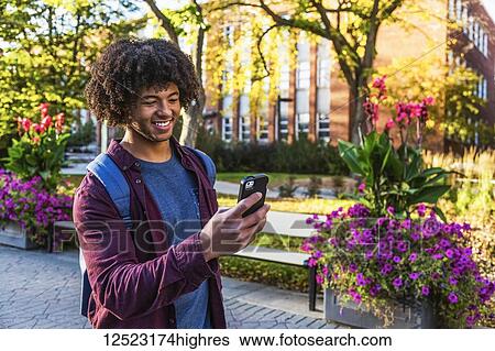 Stock Photograph - A young male African American student stands using his smart phone on a university campus; Edmonton, Alberta, Canada. Fotosearch