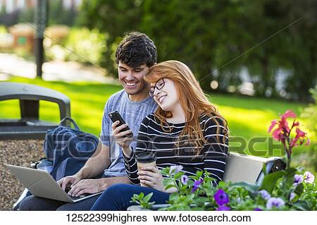 Stock Photograph - A young man and young woman sit together on a bench on the university campus using a laptop and checking social media on a smart phone while drinking coffee; Edmonton, Alberta, Canada. Fotosearch