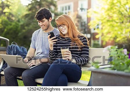 Stock Photograph - A young man and young woman sit together on a bench on the university campus using a laptop and checking social media on a smart phone while drinking coffee; Edmonton, Alberta, Canada. Fotosearch