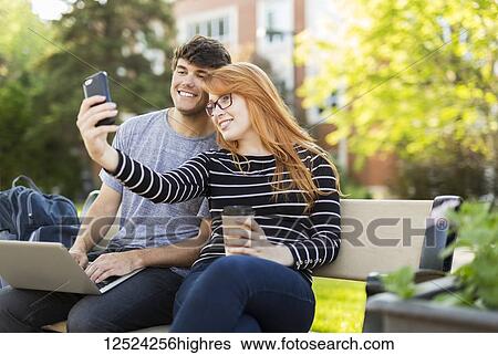 Stock Photograph - A young man and young woman sit together on a bench on the university campus taking a self-portrait on a smart phone while drinking coffee; Edmonton, Alberta, Canada. Fotosearch