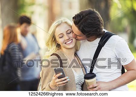 Stock Photograph - A young man kisses a young woman on the cheek while she is checking social media on a smart phone on a university campus; Edmonton, Alberta, Canada. Fotosearch