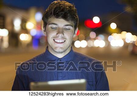 Stock Photograph - A young man uses a tablet along a street at dusk with the glowing screen illuminating his face; Edmonton, Alberta, Canada. Fotosearch
