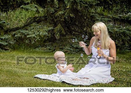 Stock Photograph - A young mom and her baby daughter in white dresses, sitting on a blanket and blowing bubbles in a park on a warm summer day; Edmonton, Alberta, Canada. Fotosearch