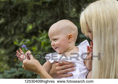 Stock Photograph - A young mom showing her baby daughter a flower while enjoying quality time together outdoors in a park on a warm summer day; Edmonton, Alberta, Canada. Fotosearch