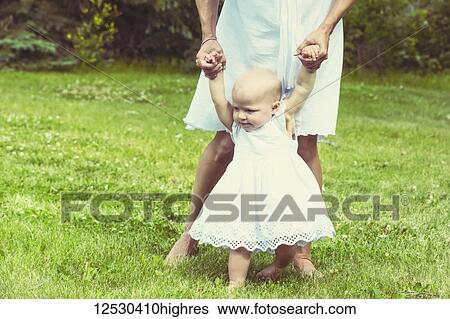 Stock Photograph - A young mom teaching her baby daughter to walk in a park on a beautiful warm sunny day; Edmonton, Alberta, Canada. Fotosearch