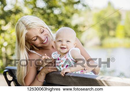Stock Photograph - A young mother spending quality time with her baby daughter and sitting on a park bench beside a manmade lake in a city park; Edmonton, Alberta, Canada. Fotosearch