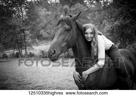 Stock Photograph - A young woman, a horse trainer and rider, sits and embraces her horse and poses for the camera; British Columbia, Canada. Fotosearch