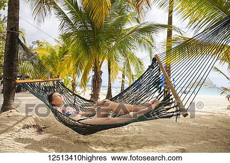 Stock Photograph - A young woman in a bikini lays in a hammock on a tropical beach with the ocean in the background; Negril, Jamaica. Fotosearch