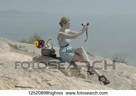 Stock Photograph - A young woman in stiletto heels sits on a rock with a picnic basket using binoculars on the coast; Spain. Fotosearch