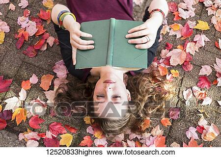 Stock Photograph - A young woman lays on her back on the ground with fallen autumn leaves reading a book; New Westminster, British Columbia, Canada. Fotosearch