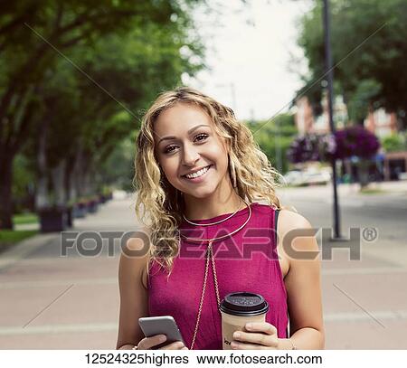 A young woman looks at the camera while walking down a street with a coffee near a university campus and texting on her smart phone; Edmonton, Alberta, Canada View Large Photo Image Stock Photograph - A young woman looks at the camera while walking down a street with a coffee near a university campus and texting on her smart phone; Edmonton, Alberta, Canada. Fotosearch