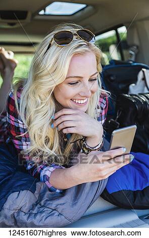 Stock Photograph - A young woman on a road trip lays in the back of a vehicle with a sleeping bag using her smart phone; Edmonton, Alberta, Canada. Fotosearch