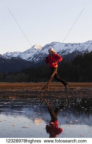 Stock Photograph - A young woman runs along the water's edge with her reflection in the water and the Kenai Mountains in the background, Kachemak Bay State Park; Alaska, United States of America. Fotosearch