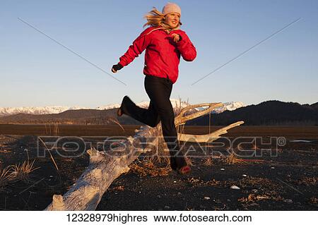 Stock Photograph - A young woman runs over pieces of driftwood with the Kenai Mountains in the background, Kachemak Bay State Park; Alaska, United States of America. Fotosearch