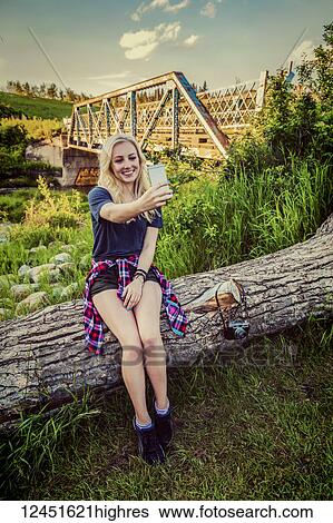 A young woman sits on a log in a park taking a self-portrait with her cell phone with a bridge and river in the background; Edmonton, Alberta, Canada View Large Photo Image Stock Photograph - A young woman sits on a log in a park taking a self-portrait with her cell phone with a bridge and river in the background; Edmonton, Alberta, Canada. Fotosearch