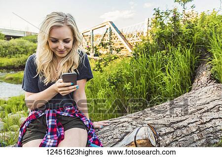 A young woman sits on log in a park using her cell phone with a river and bridge in the background; Edmonton, Alberta, Canada View Large Photo Image Stock Photograph - A young woman sits on log in a park using her cell phone with a river and bridge in the background; Edmonton, Alberta, Canada. Fotosearch