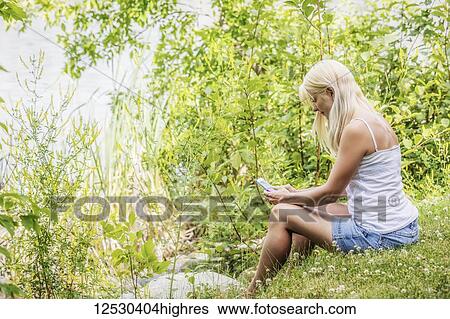 A young woman sitting on the shoreline of a man-made lake looking at her smart phone and enjoying a warm summer day: Edmonton, Alberta, Canada View Large Photo Image Stock Photograph - A young woman sitting on the shoreline of a man-made lake looking at her smart phone and enjoying a warm summer day: Edmonton, Alberta, Canada. Fotosearch