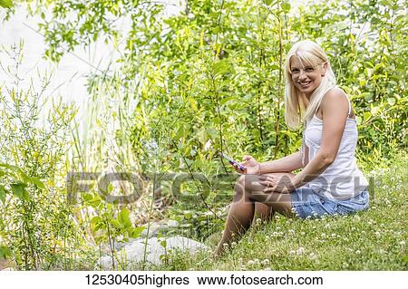 Stock Photograph - A young woman sitting on the shoreline of a man-made lake looking up and posing for the camera after using at her smart phone while enjoying a warm summer day: Edmonton, Alberta, Canada. Fotosearch