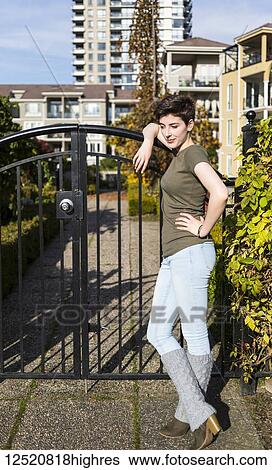 Stock Photograph - A young woman stands leaning against a metal gate looking confident with housing in the background; New Westminster, British Columbia, Canada. Fotosearch