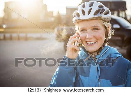 Stock Photograph - A young woman wearing a bicycle helmet is backlit by sunlight and talking on her cell phone; Anchorage, Alaska, United States of America. Fotosearch