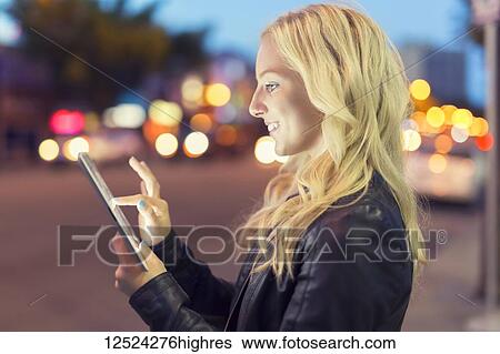 Stock Photograph - A young woman with blond hair uses a tablet along a street at dusk with the glowing screen illuminating her face; Edmonton, Alberta, Canada. Fotosearch