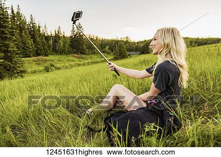 A young woman with long blond hair takes a self-portrait with a selfie stick and smart phone while sitting in a grass field in a park; Edmonton, Alberta, Canada View Large Photo Image Stock Photograph - A young woman with long blond hair takes a self-portrait with a selfie stick and smart phone while sitting in a grass field in a park; Edmonton, Alberta, Canada. Fotosearch