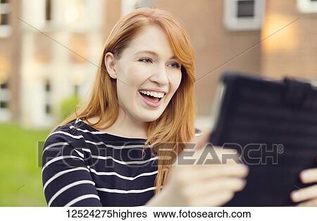 Stock Photograph - A young woman with red hair smiles big while looking at a tablet; Edmonton, Alberta, Canada. Fotosearch
