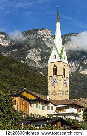 Stock Photograph - Alpine village stone church clock tower against a mountain with blue sky; Caldaro, Bolzano, Italy. Fotosearch