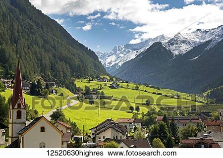Stock Photograph - Alpine village with church steeple, green meadows in the valley and snow covered mountains in the distance; St. Jodok, Tyrol, Austria. Fotosearch