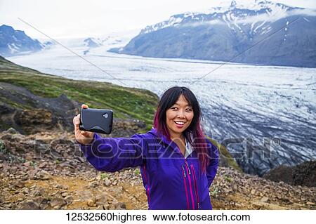 Stock Photograph - An asian female hiker takes a self-portrait from the mountain top with the glacier in the background at Vatnajokull National Park; Iceland. Fotosearch