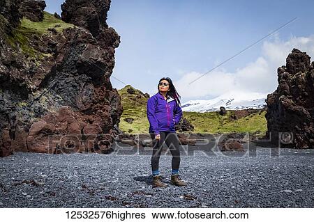 Stock Photograph - An Asian female tourist poses for a portrait on the black sand beach next to a rock formation in Western Iceland, Snaefellsnes peninsula; Iceland. Fotosearch