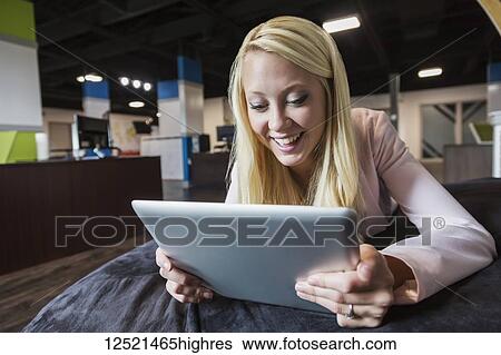 Stock Photograph - Beautiful young millennial business woman with long blond hair in the workplace on a beanbag chair using a tablet; Sherwood Park, Alberta, Canada. Fotosearch