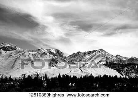 Stock Photograph - Black and white image of a snow covered mountain range under a cloudy sky with a forest in a valley; Mammoth Lakes, California, United States of America. Fotosearch
