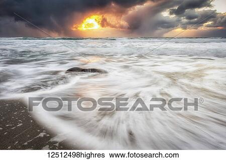 Stock Photograph - Blur of the tide washing up on the sandy shore along the coast and a golden sun peaking through storm clouds; Nordland, Norway. Fotosearch