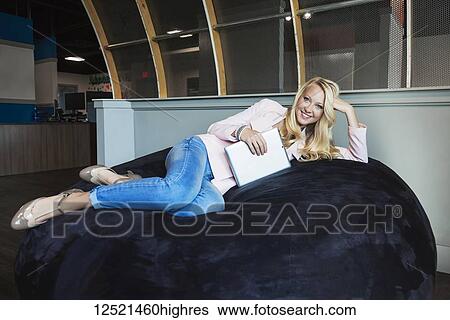 Stock Photograph - Business portrait of a beautiful young millennial businesswoman with long blond hair holding a tablet and posing for the camera on a beanbag chair in the workplace; Sherwood Park, Alberta, Canada. Fotosearch