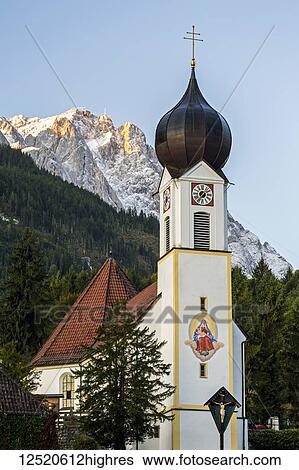 Stock Photograph - Church with painted fresco and clock tower against a glowing mountain background at sunrise with blue sky; Grainau, Bavaria, Germany. Fotosearch