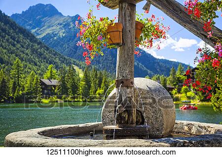Stock Photograph - Close-up of a stone fountain with flower pots by Champex Lake and mountain range in the background; Champex, Valais, Switzerland. Fotosearch