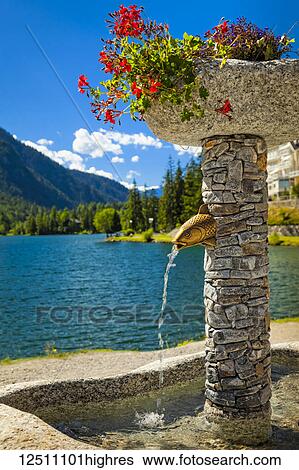 Stock Photograph - Close-up of a stone fountain with flower pots by Champex Lake and mountain range in the background; Champex, Valais, Switzerland. Fotosearch