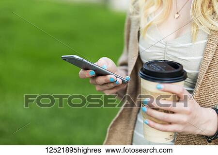 Stock Photograph - Close-up of a young woman's hands with blue fingernail polish holding a coffee cup and texting on her smart phone; Edmonton, Alberta, Canada. Fotosearch