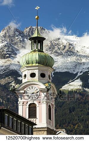 Stock Photograph - Decorative church clock tower with forest and rugged, snow covered peaks and blue sky in background; Innsbruck, Tyrol, Austria. Fotosearch
