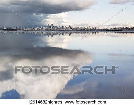 Stock Photograph - Distant view of the Vancouver skyline over the Pacific Ocean reflecting clouds, viewed from Spanish Banks; Vancouver, British Columbia, Canada. Fotosearch