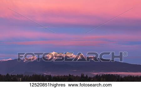 Stock Photograph - Dramatic glowing clouds above the snow-capped mountains and reflecting on the snow below; British Columbia, Canada. Fotosearch