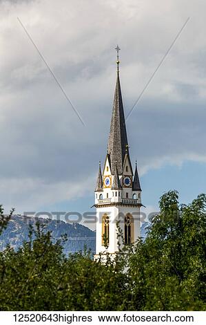 Stock Photograph - Elaborate church clock tower with trees in foreground and dark clouds in the background; Lienz, Tyrol, Austria. Fotosearch